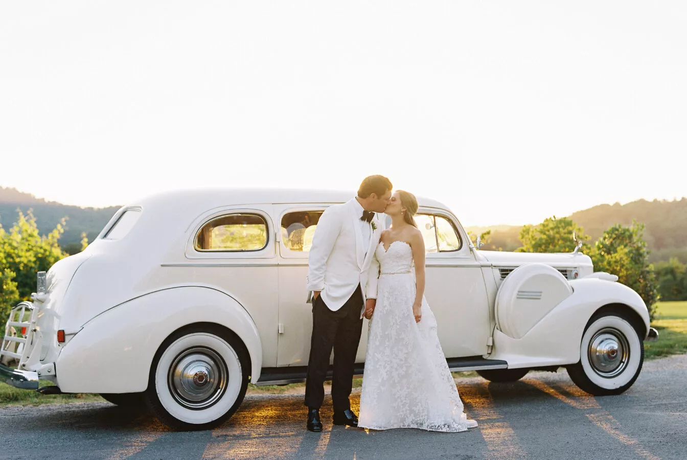 A bride and groom kiss in front of a classic white wedding car